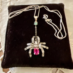 May include: A silver necklace with a spider pendant. The pendant has a large, oval, pink gemstone at its centre, surrounded by smaller clear stones. The spider's body is detailed with clear stones and the necklace is displayed on a black velvet surface.