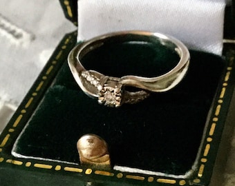 Könnte beinhalten: Ein silberner Ring mit einem einzelnen Diamanten in der Mitte und kleineren Diamanten auf der Ringschiene. Der Ring befindet sich in einer grünen Samt-Ringbox.