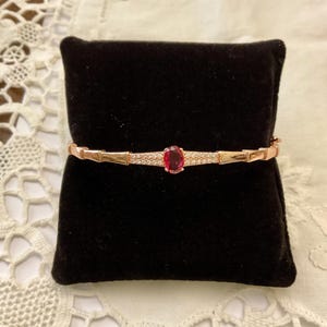 Könnte beinhalten: Roségoldenes Armband mit einem ovalen Rubin in der Mitte und einem Band aus kleinen, funkelnden Steinen. Das Armband wird auf einem schwarzen Samtkissen präsentiert, das sein elegantes Design hervorhebt.
