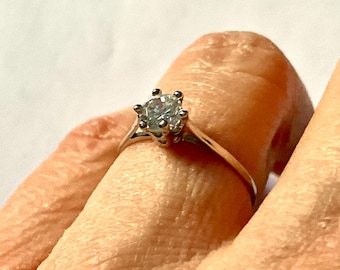 Könnte beinhalten: Ein silberner Verlobungsring mit einem großen, klaren, rund geschliffenen Edelstein. Der Ring hat eine klassische Sechs-Zacken-Fassung, und das Band ist dünn und poliert. Der Ring wird an einem Finger gezeigt.