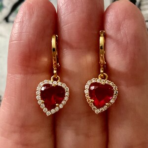 Peut inclure: Une paire de boucles d'oreilles créoles dorées avec des pierres centrales rouges en forme de cœur. Chaque cœur est entouré d'un halo de petites pierres claires et scintillantes. Les boucles d'oreilles ont un fermoir à charnière en haut.