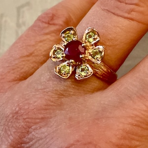 Peut inclure: Une bague en or avec une pierre précieuse rouge au centre entourée de petites pierres précieuses vertes, en forme de fleur.