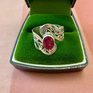 Op de afbeelding: Een zilveren ring met een grote ovale robijn edelsteen. De ring heeft een open ontwerp met decoratieve krullen en wordt gepresenteerd in een groen fluwelen doosje. De ring is een sieraad.