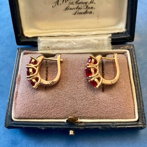 Peut inclure: Paire de boucles d'oreilles dorées à fermoir créole, chacune ornée de trois pierres rouges, présentées dans un écrin à bijoux vintage bleu foncé avec un intérieur rose. Le texte "Jewellers, A. Woodhouse & Son Ltd" est visible.