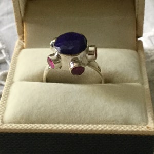 Peut inclure: Une bague en argent avec une grande pierre précieuse bleue entourée de quatre petites pierres précieuses roses.