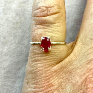 Pode incluir: Um anel de prata com uma vibrante pedra preciosa vermelha oval. O anel é exibido em um dedo, destacando a cor da pedra preciosa e o design simples e elegante do anel. A pedra preciosa é o ponto focal.