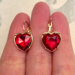 Peut inclure: Une paire de boucles d'oreilles en forme de cœur avec un cadre doré et une grosse pierre précieuse rouge facettée. Les boucles d'oreilles ont une fermeture à levier. Les bijoux sont présentés sur un fond neutre.