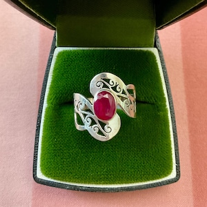 May include: A silver ring featuring an oval, deep red gemstone, set within an openwork design. The ring is presented in a green velvet-lined box with a black border, against a pink backdrop.