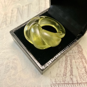 May include: A light green, knotted ring displayed in a black jewellery box with a velvet lining. The ring is translucent with a smooth, rounded shape and a subtle yellow tint. The box has a shiny, dark exterior.