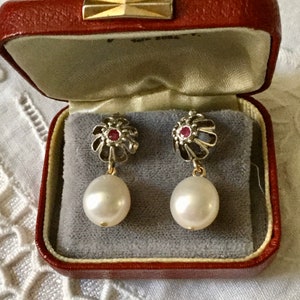 May include: A pair of vintage drop earrings in a red jewellery box. The earrings have a silver-toned floral design with a red gemstone centre and a large, white pearl drop. The box has a gold clasp.