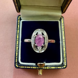 Peut inclure: Bague vintage avec une pierre rose de forme ovale, ornée de petits diamants. La bague est présentée dans un écrin à bijoux doublé de velours bleu avec un fermoir doré, sur fond rose.