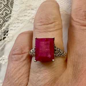 Peut inclure: Une bague en argent ornée d'une grosse pierre rouge foncé de forme rectangulaire. La bague présente un motif filigrané orné sur le corps. La pierre est sertie de quatre griffes.