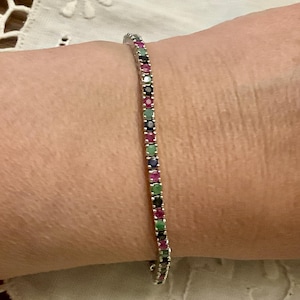 Peut inclure: Un bracelet en argent orné d'une rangée de pierres précieuses colorées. Les pierres sont un mélange de rouge, vert, bleu et noir. Le bracelet est porté au poignet.