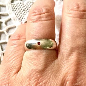 Peut inclure: Une bague argentée avec une petite pierre précieuse rouge ronde au centre. La bague est épaisse et lisse, avec une finition polie. La bague est sur un doigt.