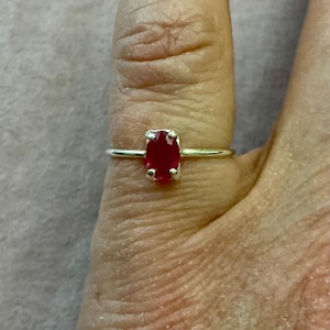 Op de afbeelding: Een zilveren ring met een levendige rode ovale edelsteen. De edelsteen is gezet in een vier-klauwzetting en de band is dun en delicaat. De ring wordt op een vinger getoond.