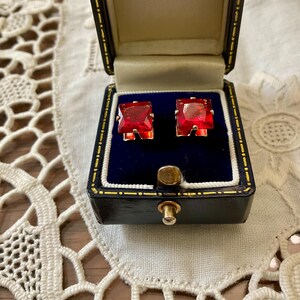 Peut inclure: Une paire de boucles d'oreilles avec des pierres précieuses rouges de taille carrée serties dans du métal doré, présentées dans un écrin à bijoux doublé de velours bleu foncé avec un extérieur noir et un fermoir en métal doré.