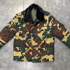 May include: Camouflage pattern jacket with a black faux fur collar. The jacket features a button-up front, multiple pockets, and a green, brown, and tan color scheme. The jacket is laid flat on a gray surface.