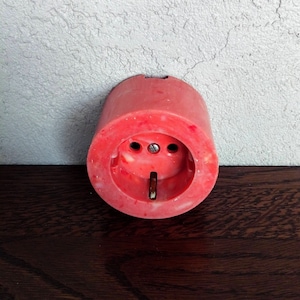 Puede incluir: Una toma de corriente eléctrica de plástico rojo con dos agujeros redondos y una sola ranura. La toma está montada en una pared blanca.