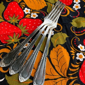 May include: Four silver forks with decorative handles are arranged on a black fabric with a strawberry and floral pattern. The fabric features large red strawberries, orange leaves, and white flowers.