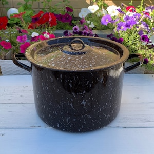 May include: A black enamel pot with a white speckled pattern and a lid. The pot has two handles and is sitting on a white wooden surface.
