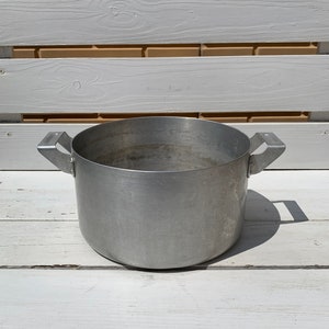 Peut inclure: Une grande casserole en aluminium argentée avec deux poignées. La casserole est vide et repose sur une surface en bois blanc.
