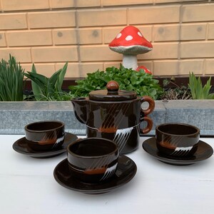 Puede incluir: Una tetera de cerámica marrón y blanca con un juego a juego de cuatro tazas y platillos. La tetera tiene un diseño único con rayas marrones y acentos blancos. Las tazas y los platillos tienen un diseño similar.