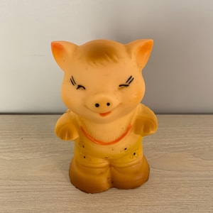 Op de afbeelding: Een geel rubberen varkenspeelgoed met een bruin gezicht, gesloten ogen en een kleine glimlach. Het varken draagt een geel en oranje outfit met zwarte stippen.
