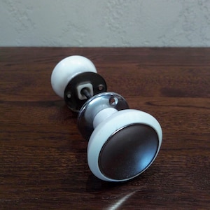 Puede incluir: Pomo de puerta de porcelana blanca con base de metal plateado y centro gris oscuro.
