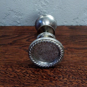 Peut inclure: Une poignée de porte argentée avec un motif de corde autour du bord. La poignée est sur une surface en bois.