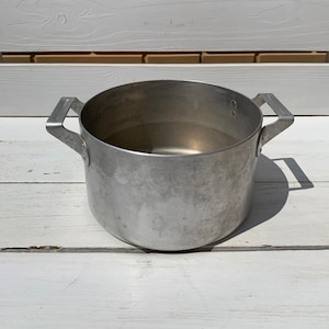 May include: A silver aluminum pot with two handles. The pot is empty and sitting on a white wooden surface.