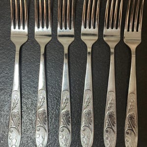 May include: Six silver forks with ornate floral and leaf designs on the handles. The tines are evenly spaced. The forks are arranged in a row on a dark surface, showcasing a classic silverware set.