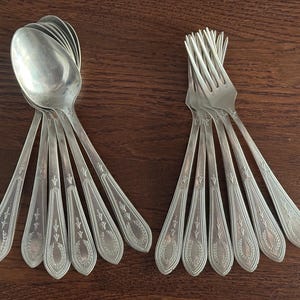 May include: A collection of vintage silverware, including spoons and forks, arranged on a dark wooden surface. The spoons and forks are silver-toned, with decorative detailing on the handles. The spoons are stacked together, while the forks are grouped separately.