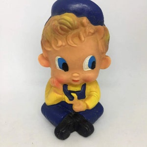 Op de afbeelding: Een vintage rubberen speelgoed van een jongen die een blauwe pet, een geel shirt en een blauwe overall draagt. Het speelgoed heeft een lieve uitdrukking en zit met zijn handen in elkaar.
