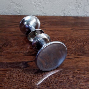 May include: A silver door knob with a round, flat base and a decorative ring around the center.