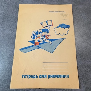 May include: A vintage drawing notebook with a yellow cover and a blue and red illustration of two children flying a paper airplane. The text "Тетрадь для рисования" is printed in black on the cover.