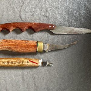 Könnte beinhalten: Drei Messer mit Holzgriffen. Das oberste Messer hat einen Griff mit Fingermulden und eine gerade Klinge. Das mittlere Messer besitzt eine gebogene Klinge und eine Messingzwinge. Das untere Werkzeug hat einen strukturierten Holzgriff.