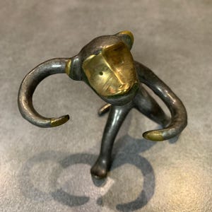 Puede incluir: Una figurita de mono de bronce estilizada con la cara dorada. El mono está de pie sobre dos piernas y tiene los brazos levantados en el aire.