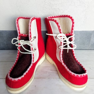 Könnte beinhalten: Ein Paar rote Filzstiefel mit weißen Verzierungen und braunen Filzdetails. Die Stiefel haben weiße Schnürsenkel und eine weiße Sohle.