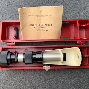 May include: A vintage microscope in a red case. The microscope is black and silver, with a white eyepiece. The case is open, revealing the instrument and a paper document with Cyrillic text. The case is red.
