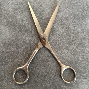 Könnte beinhalten: Eine vintage Metallschere mit verwitterter, silberfarbener Oberfläche und sichtbaren Oxidationsspuren um den zentralen Drehpunkt. Das Werkzeug verfügt über zwei symmetrische Fingerringe und spitz zulaufende Klingen in gekreuzter Position.