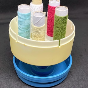 May include: A white plastic thread holder with a blue base. The holder has six slots for spools of thread. The spools are in various colors, including white, light blue, green, and red.