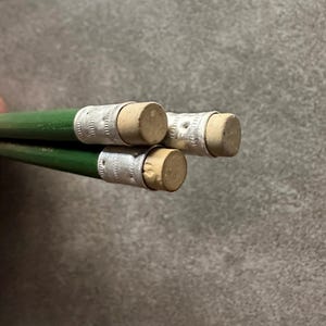 Puede incluir: Primer plano de tres lápices verdes con bandas metálicas plateadas y gomas de borrar color beige. Los lápices están en ángulo, mostrando las gomas y los cuerpos de madera verde. El fondo es gris neutro.