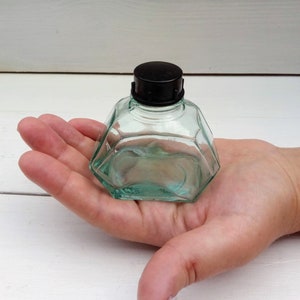 Puede incluir: Una pequeña botella de vidrio transparente con una tapa de plástico negro. La botella tiene una forma octogonal facetada.