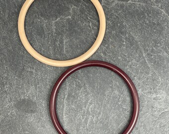 May include: Two circular bangles. One is a light tan color, and the other is a deep burgundy. The bangles are made of a smooth material and are displayed on a dark gray surface. These accessories are suitable for everyday wear.