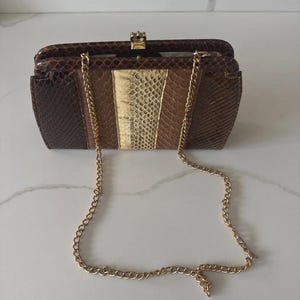 Pode incluir: Uma clutch vintage marrom e dourada com uma alça de corrente dourada. A bolsa tem formato retangular com fecho de pressão e um detalhe dourado decorativo. A frente da bolsa apresenta um padrão listrado com tons variados de marrom e dourado.