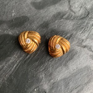 May include: A pair of gold-coloured earrings with a woven pattern. Each earring has a central, round, clear gemstone. The earrings are triangular and sit on a dark, textured surface.