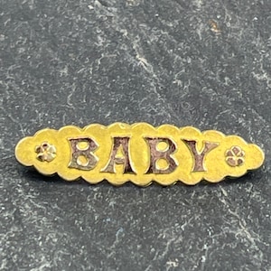 以下が含まれることがあります： 暗い文字で「BABY」という文字が入った金色のピン。ピンはスカラップエッジで、両端に小さな花の模様があります。ピンは暗く、質感のある背景に配置されています。