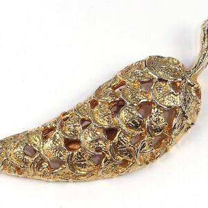 May include: A gold-colored chili pepper-shaped brooch. The brooch has a textured surface with a detailed leaf pattern. The stem of the pepper is also gold and textured. The brooch is likely made of metal and is a decorative accessory.