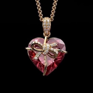 Könnte beinhalten: Eine goldfarbene Halskette mit einem herzförmigen Anhänger. Das Herz ist ein facettierter, rosa Edelstein, verziert mit einer goldenen Schleife, die mit kleinen, funkelnden Steinen besetzt ist. Die Kette ist ein filigranes, verbundenes Design, der Hintergrund ist schwarz.