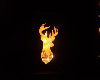 May include: A deer head silhouette with illuminated fairy lights, creating a warm, golden glow against a dark background. The decorative piece is likely for holiday or seasonal decor.